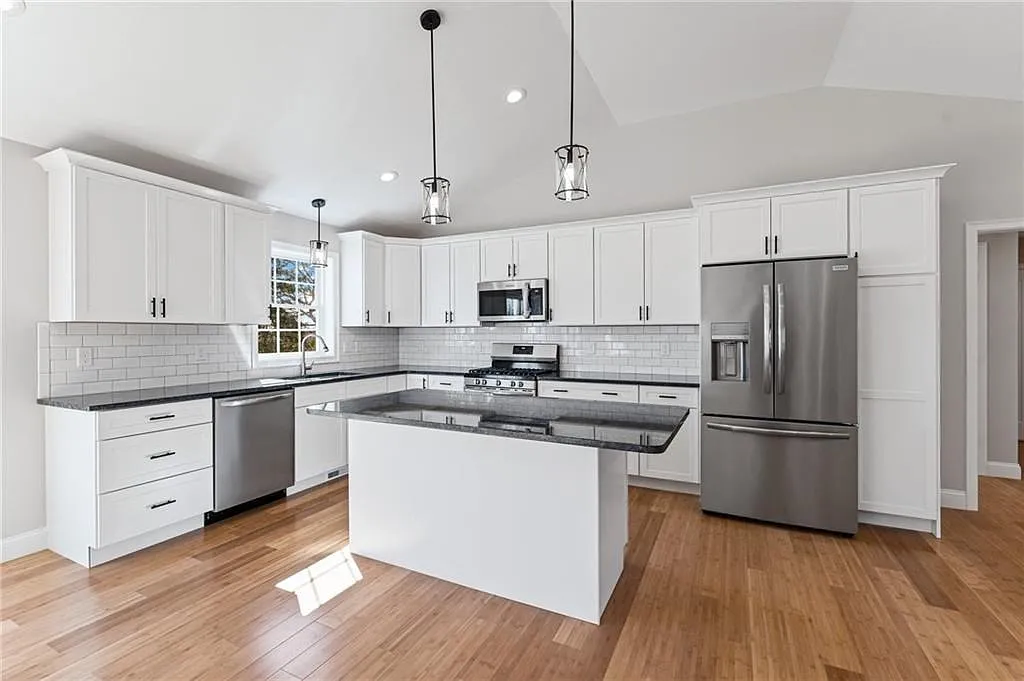Open white kitchen with subway tile, dark stone countertops, vaulted ceiling, and hardwood floors